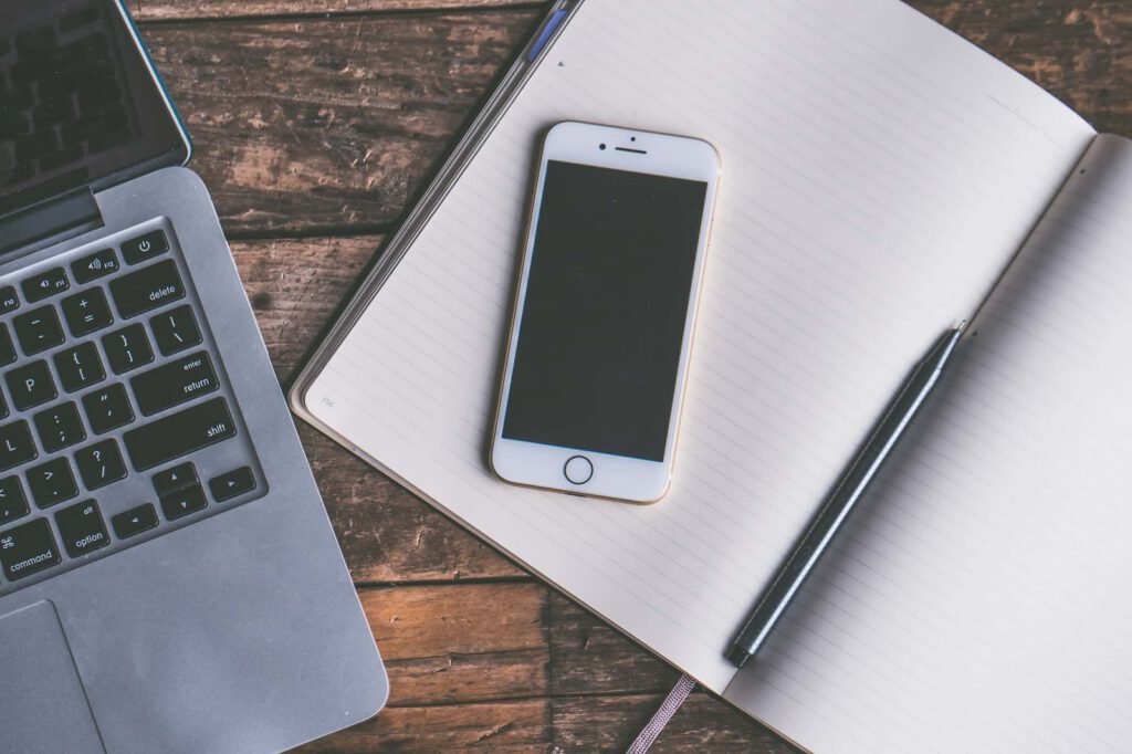 A smartphone rests on an open lined notebook with a pen, next to a laptop on a wooden surface, suggesting seamless access to a responsive website.