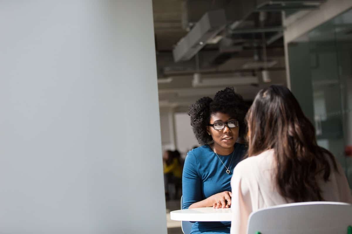 Two women sit across from each other at a table in a modern office, engaged in conversation. One faces the camera while the other has her back turned, possibly preparing to Ask Your Web Designer about creative project ideas.