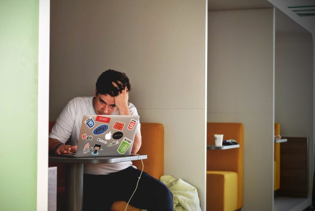 A person sits alone at a table in a modern workspace booth, focused on a laptop covered in tech stickers, working hard to achieve a faster website, with their hand resting on their forehead.