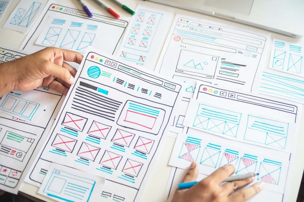 Several hand-drawn website wireframes are spread out on a table, as a person arranges and reviews the mockup layouts—demonstrating practical steps in how to design a website for higher conversion rates.