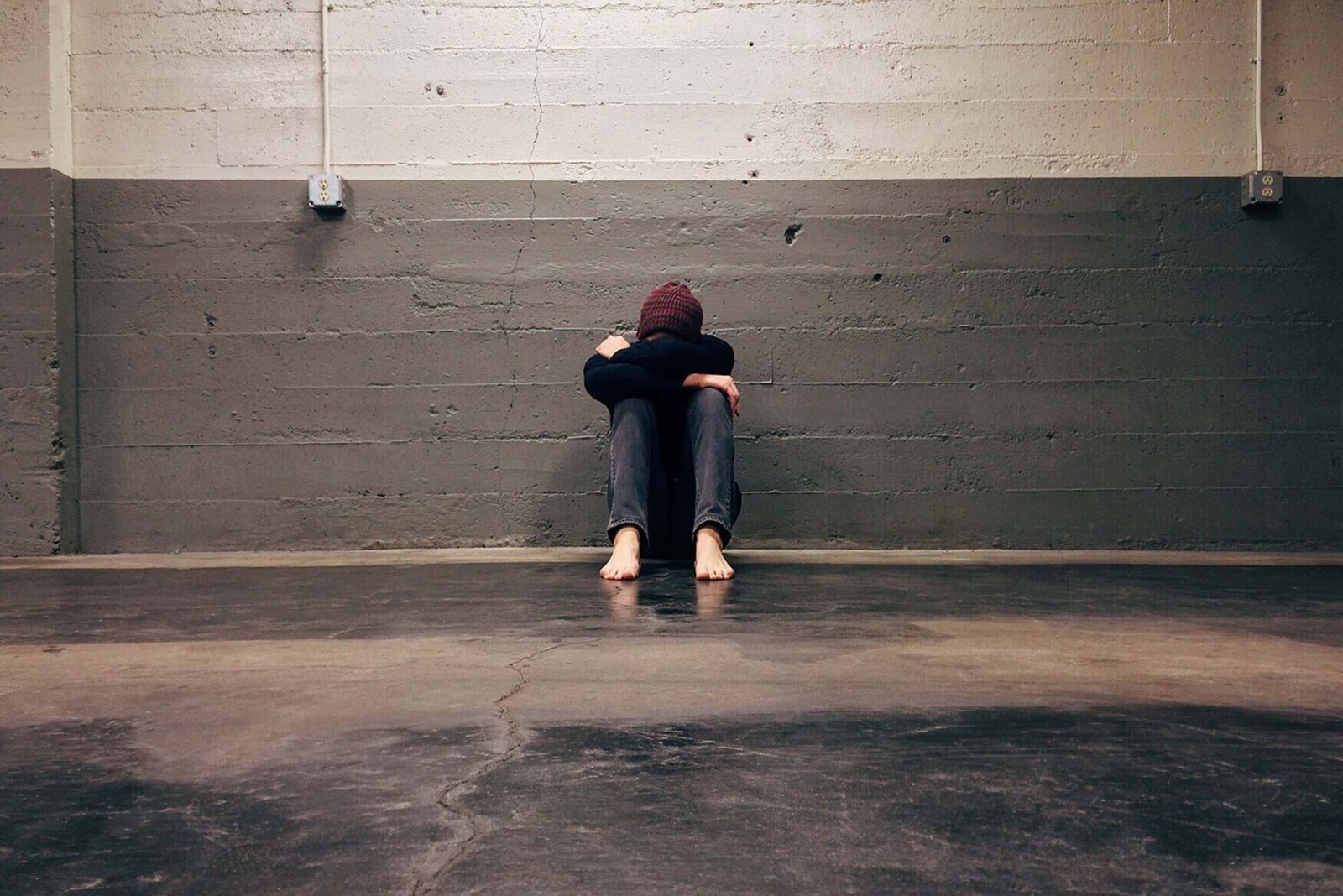 A person sits barefoot on the floor in an empty room, leaning against a gray wall with knees drawn up and head resting on crossed arms, a posture that quietly reflects anxiety.