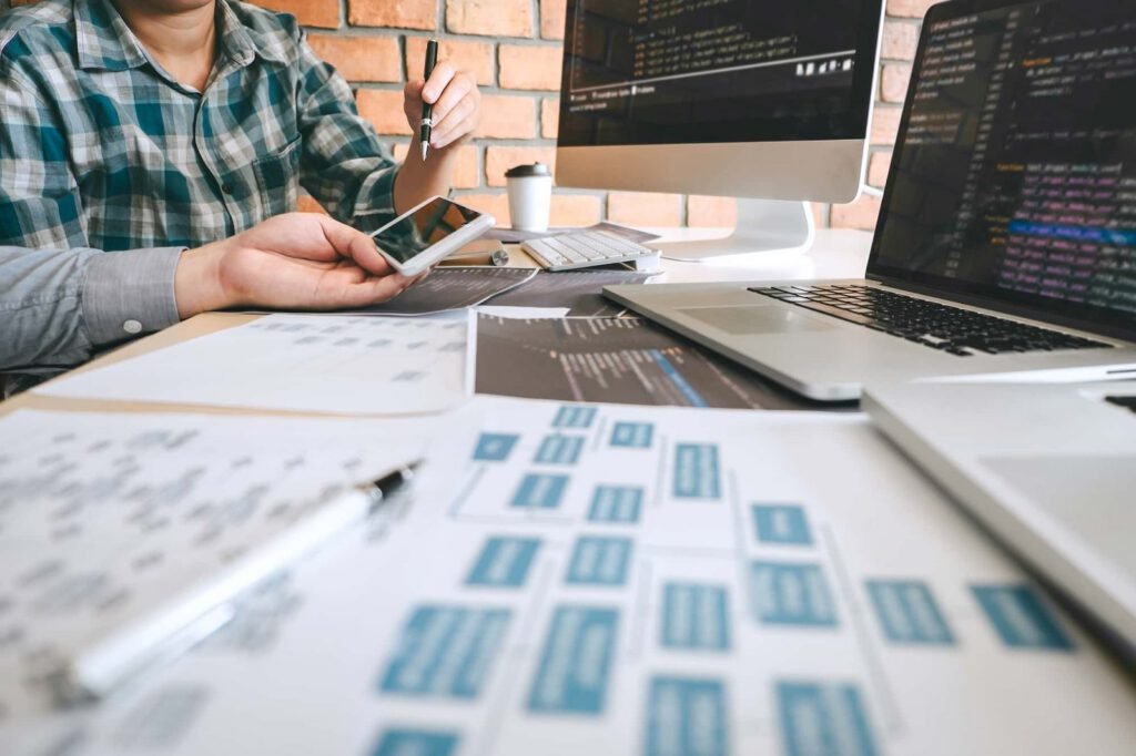 Two people discuss web design project plans at a desk with laptops, papers, and a smartphone, with coding and diagrams visible on screens and documents.