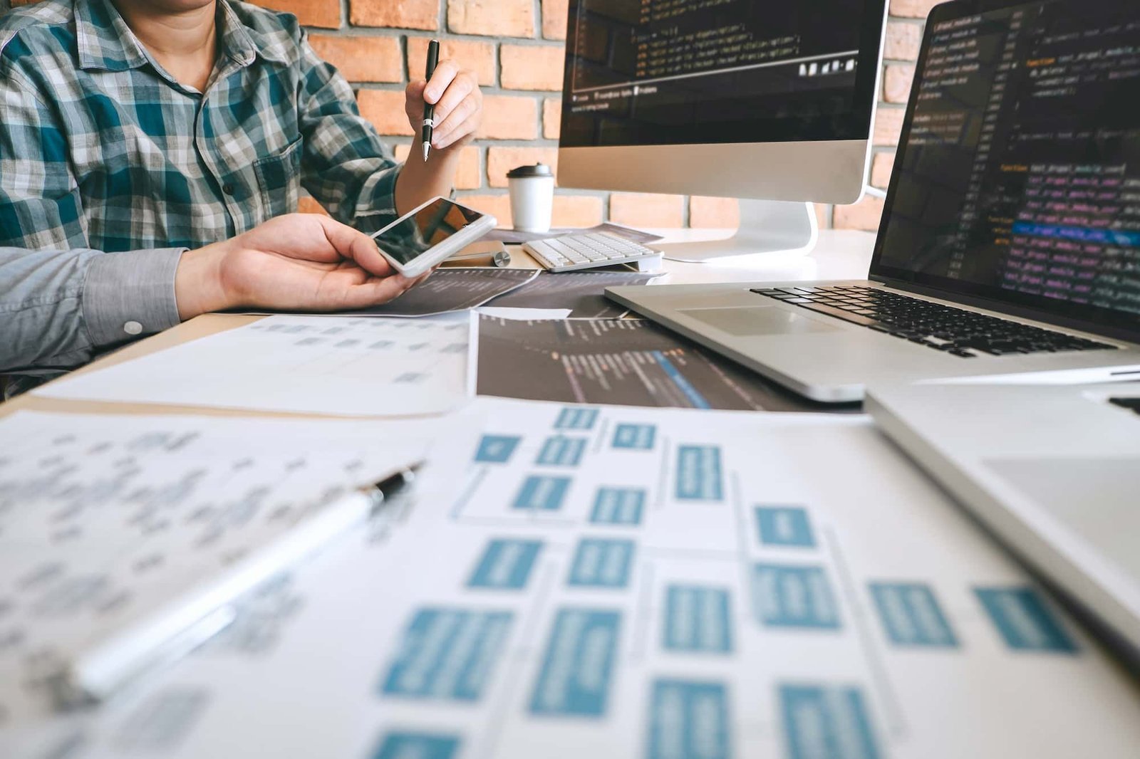Two people discuss web design project plans at a desk with laptops, papers, and a smartphone, with coding and diagrams visible on screens and documents.