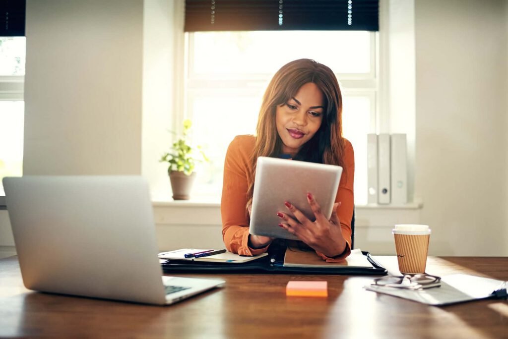 A woman sits at a desk in an office, looking at a tablet as she researches SEO Website Trends. A laptop, notebook, coffee cup, and documents are on the desk.