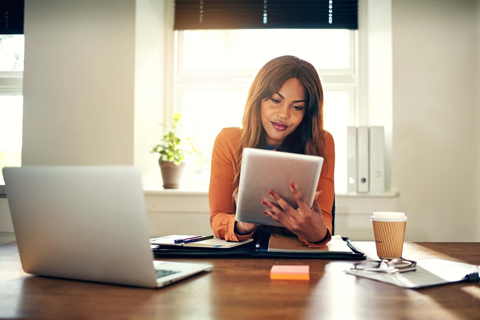 A woman sits at a desk in an office, looking at a tablet as she researches SEO Website Trends. A laptop, notebook, coffee cup, and documents are on the desk.