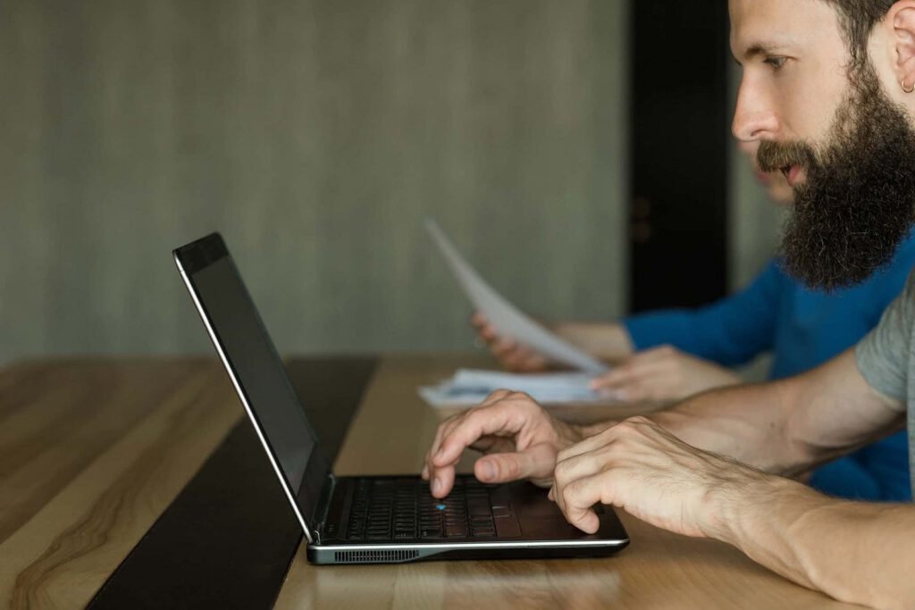 Two men sitting at a wooden table; the man in front is typing on his laptop, focused on SEO tasks, while the person in the background holds sheets of paper.