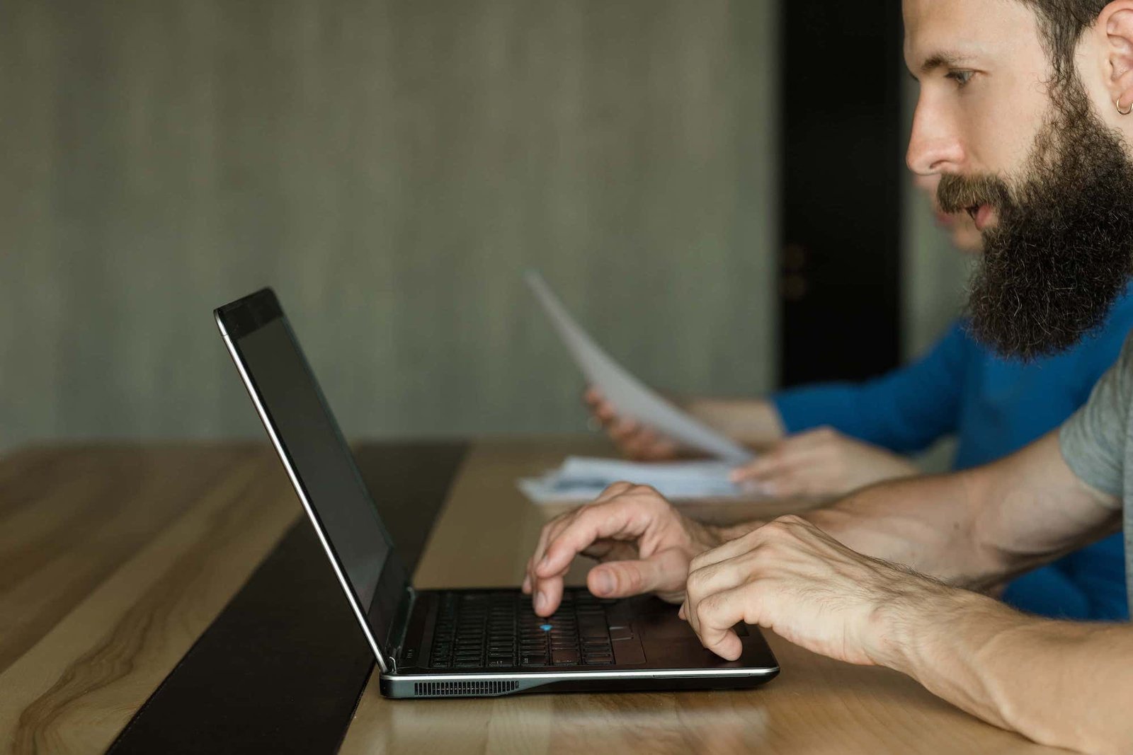 Two men sitting at a wooden table; the man in front is typing on his laptop, focused on SEO tasks, while the person in the background holds sheets of paper.