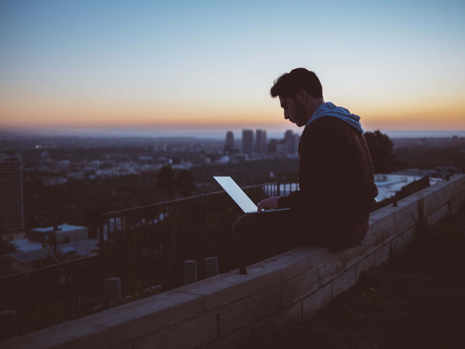 A person sits on a low wall outdoors at sunset, working on a laptop and finding balance between work and relaxation, with a city skyline visible in the background.