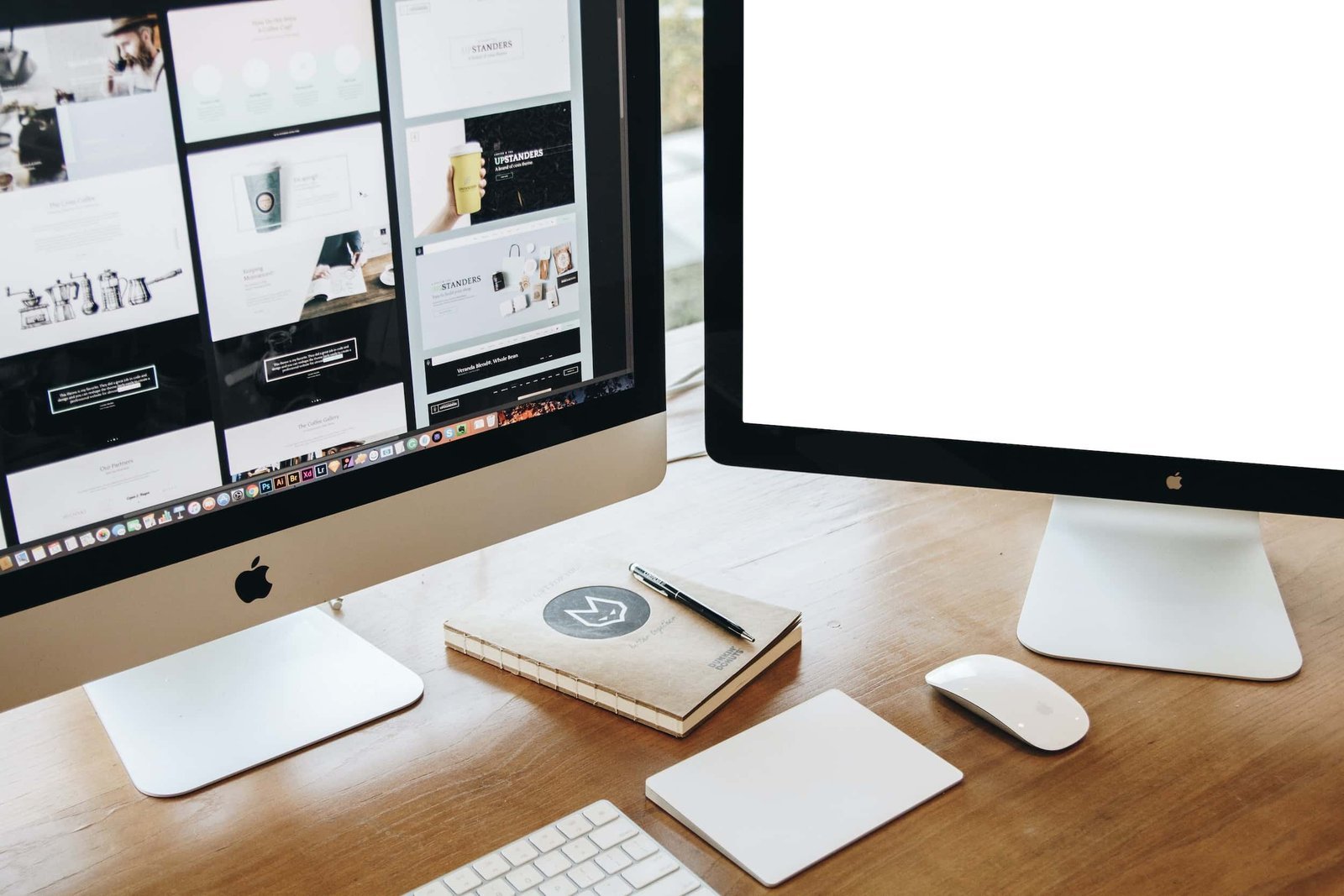 Two Apple desktop monitors on a wooden desk; one displays web design thumbnails, the other is blank. A notebook, pen, mouse, and keyboard are also visible.