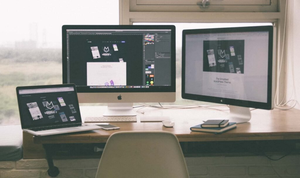 A desk set up for designing with a laptop and two large monitors displaying website designs, plus a smartphone, notebooks, mouse, and keyboard arranged neatly in front of a window.