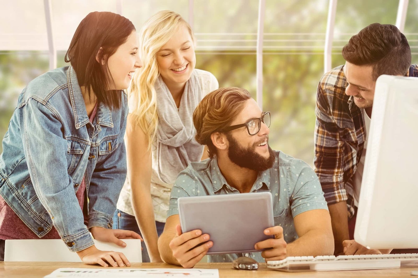 Four young adults gather around a desk, smiling and looking at each other; one person holds a tablet displaying SEO analytics, while another sits at a computer.