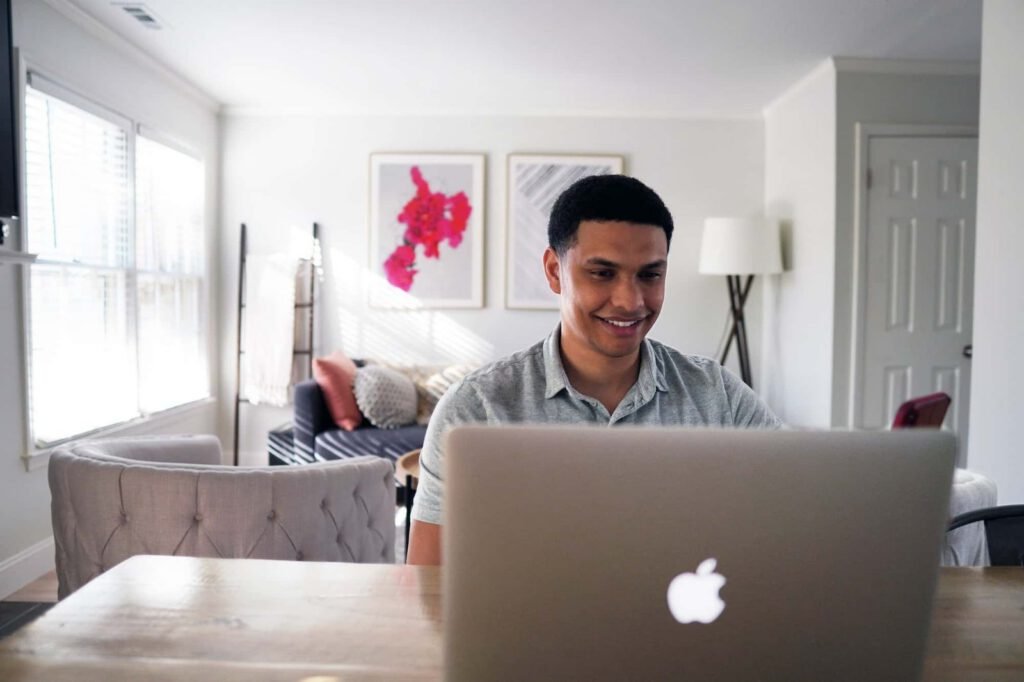 A man sits at a wooden table using a laptop, focused on Conversion Rate Optimization, in a bright, modern living room with artwork, a couch, and a floor lamp in the background.