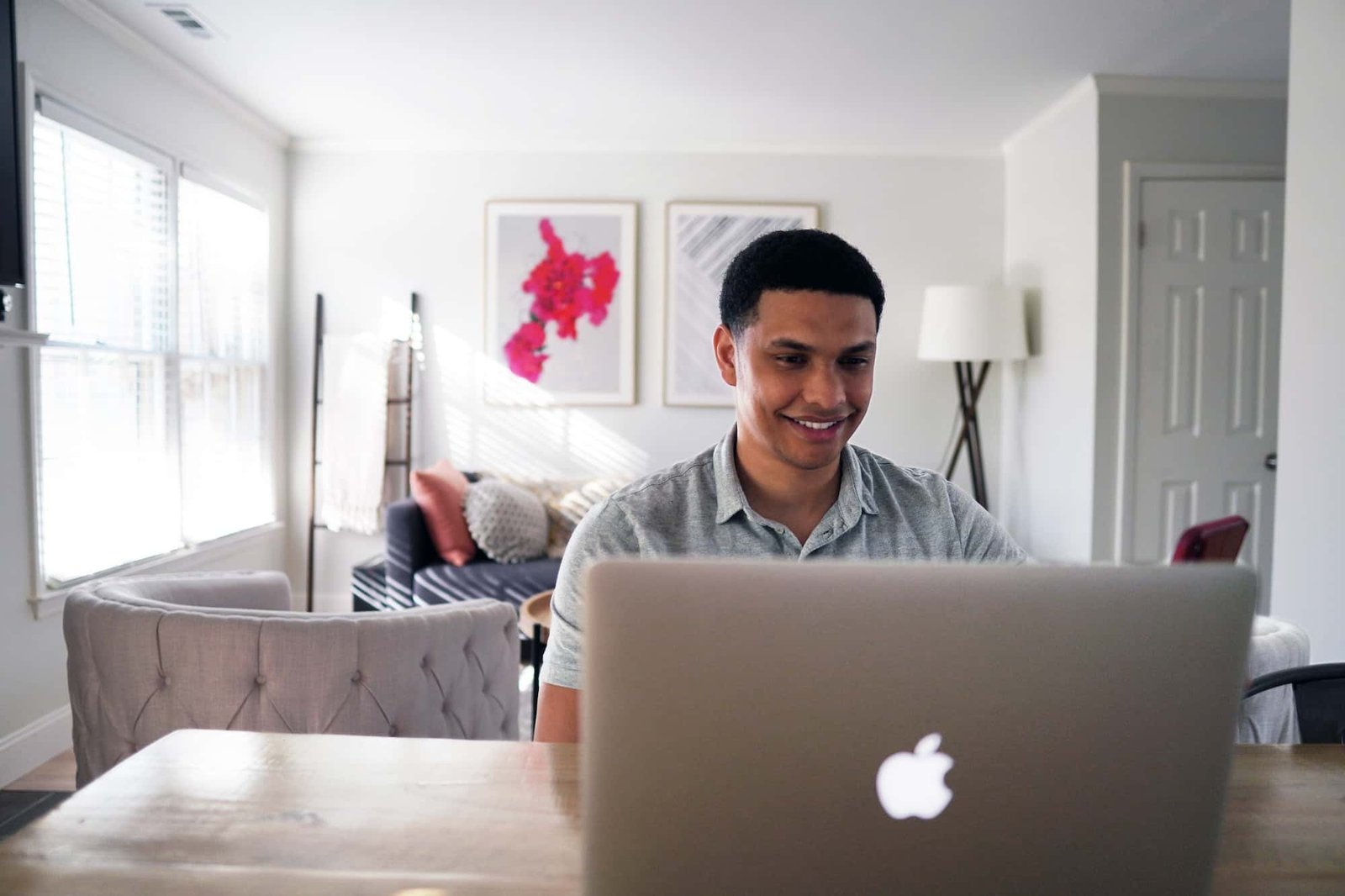 A man sits at a wooden table using a laptop, focused on Conversion Rate Optimization, in a bright, modern living room with artwork, a couch, and a floor lamp in the background.