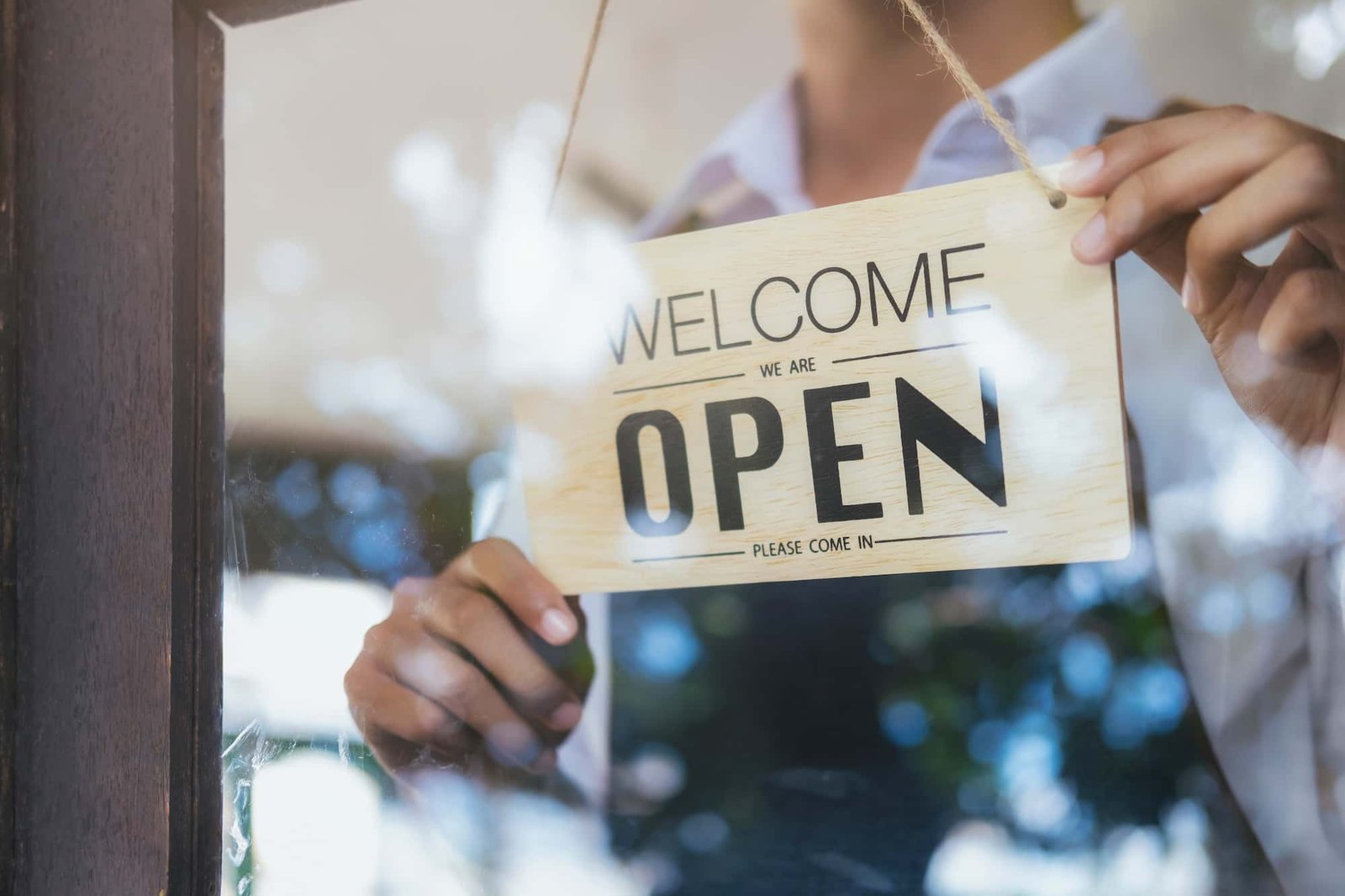 A person hangs a wooden "Welcome, We Are Open" sign on a glass door, inviting customers in and boosting Local SEO by signaling that the business is open to the community.