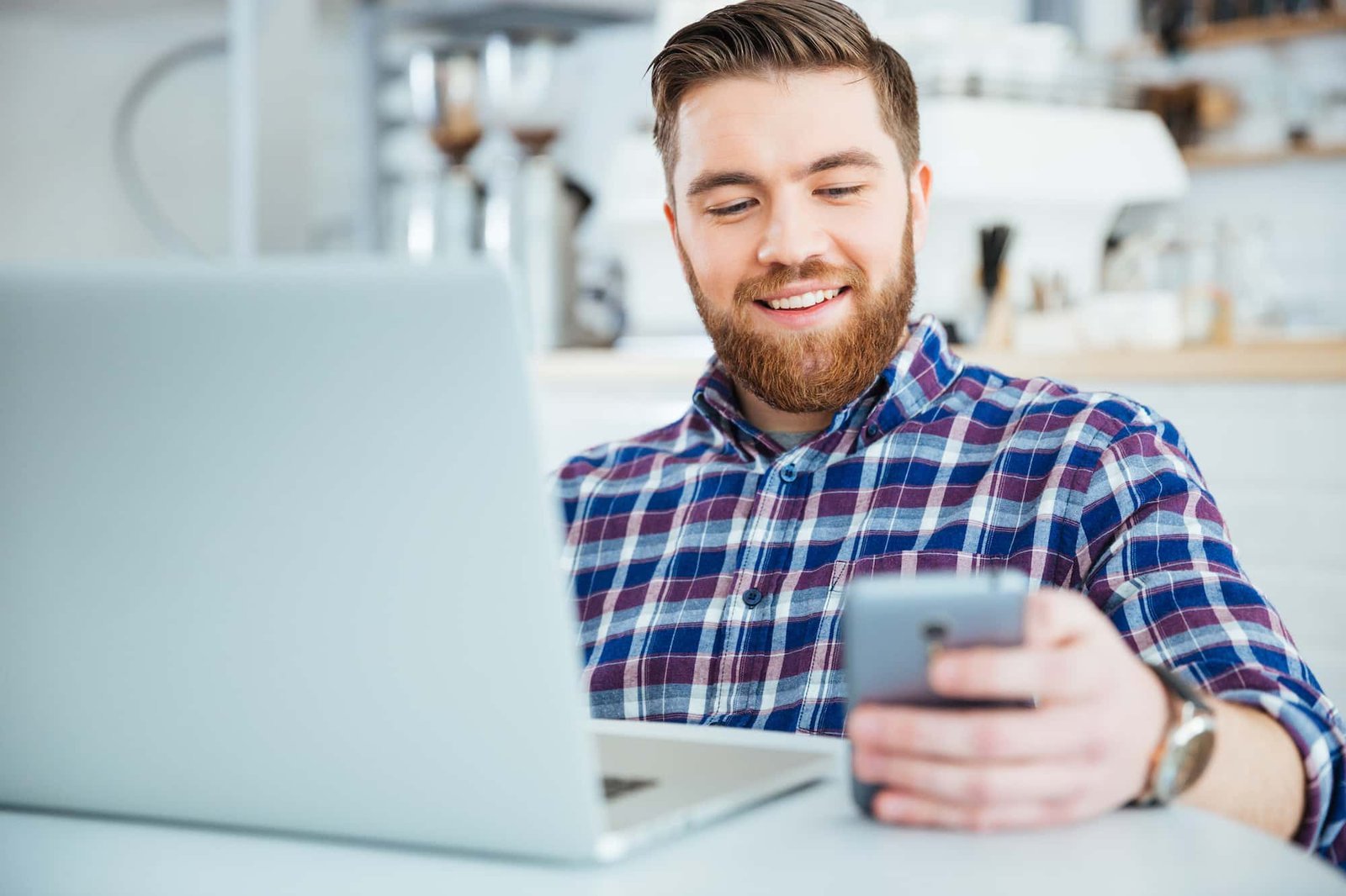 A man sits at a desk with a laptop, smiling while looking at his smartphone in a bright indoor setting—perhaps checking the latest CRO results.