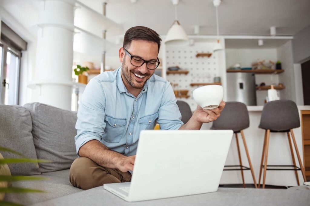 A man sits on a couch, smiling at his laptop while holding a white mug, likely reviewing CRO reports, with a kitchen visible in the background.
