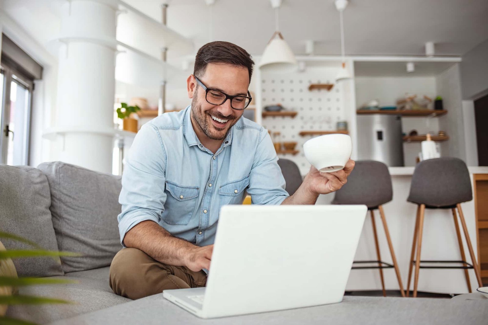 A man sits on a couch, smiling at his laptop while holding a white mug, likely reviewing CRO reports, with a kitchen visible in the background.