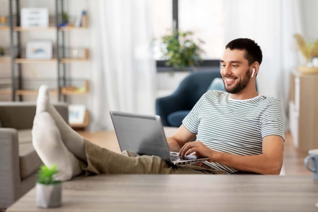 A man sits with his feet up on a table, smiling while handling maintenance tasks on his laptop and wearing wireless earbuds in a bright living room.