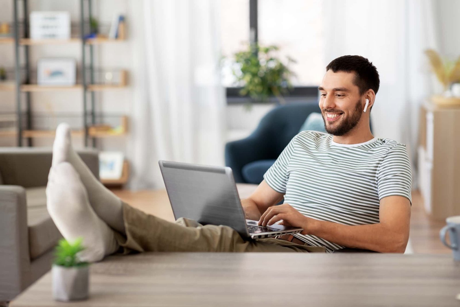 A man sits with his feet up on a table, smiling while handling maintenance tasks on his laptop and wearing wireless earbuds in a bright living room.