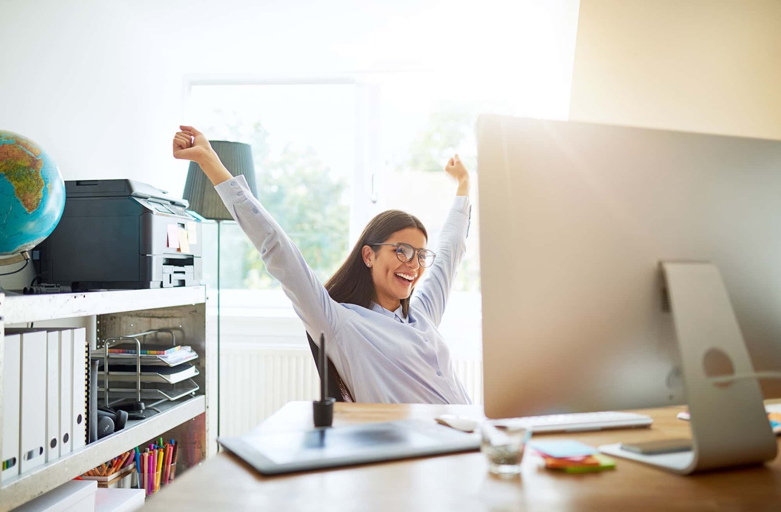A woman wearing glasses stretches and smiles at her desk in a bright office, working on SEO tasks with a computer, printer, and shelves in the background.