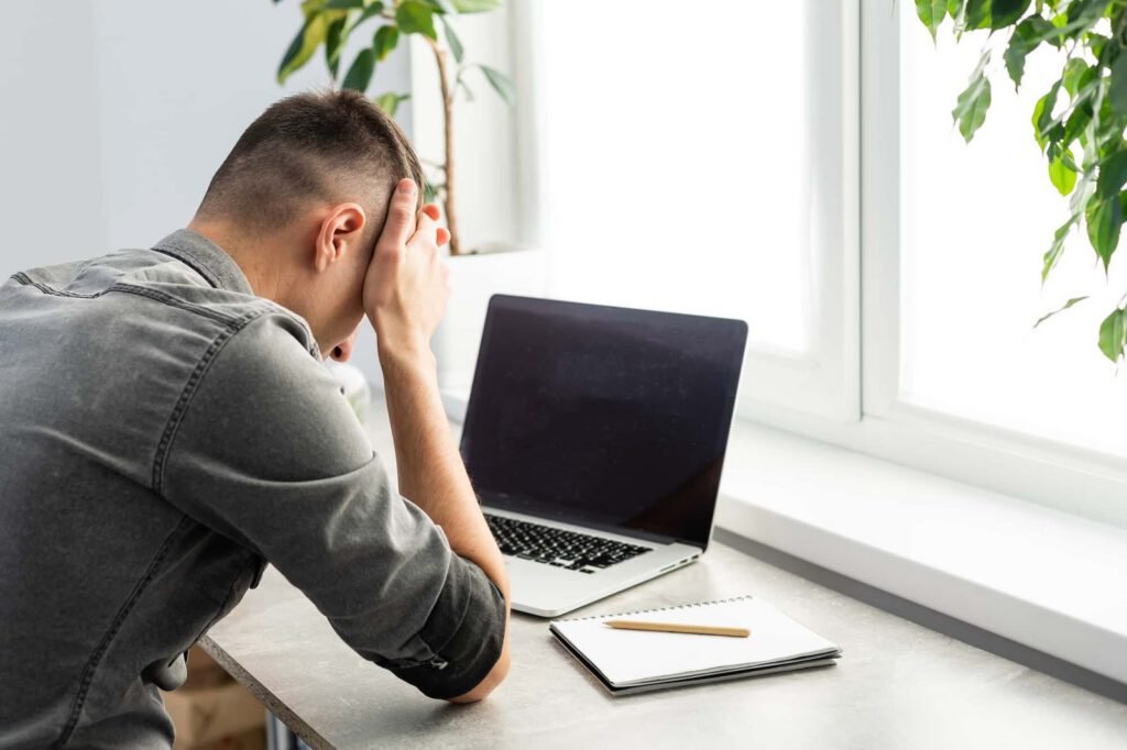 A person sits at a desk with their head in their hands in front of a closed laptop and an open notebook, appearing stressed or frustrated by unexpected maintenance issues.