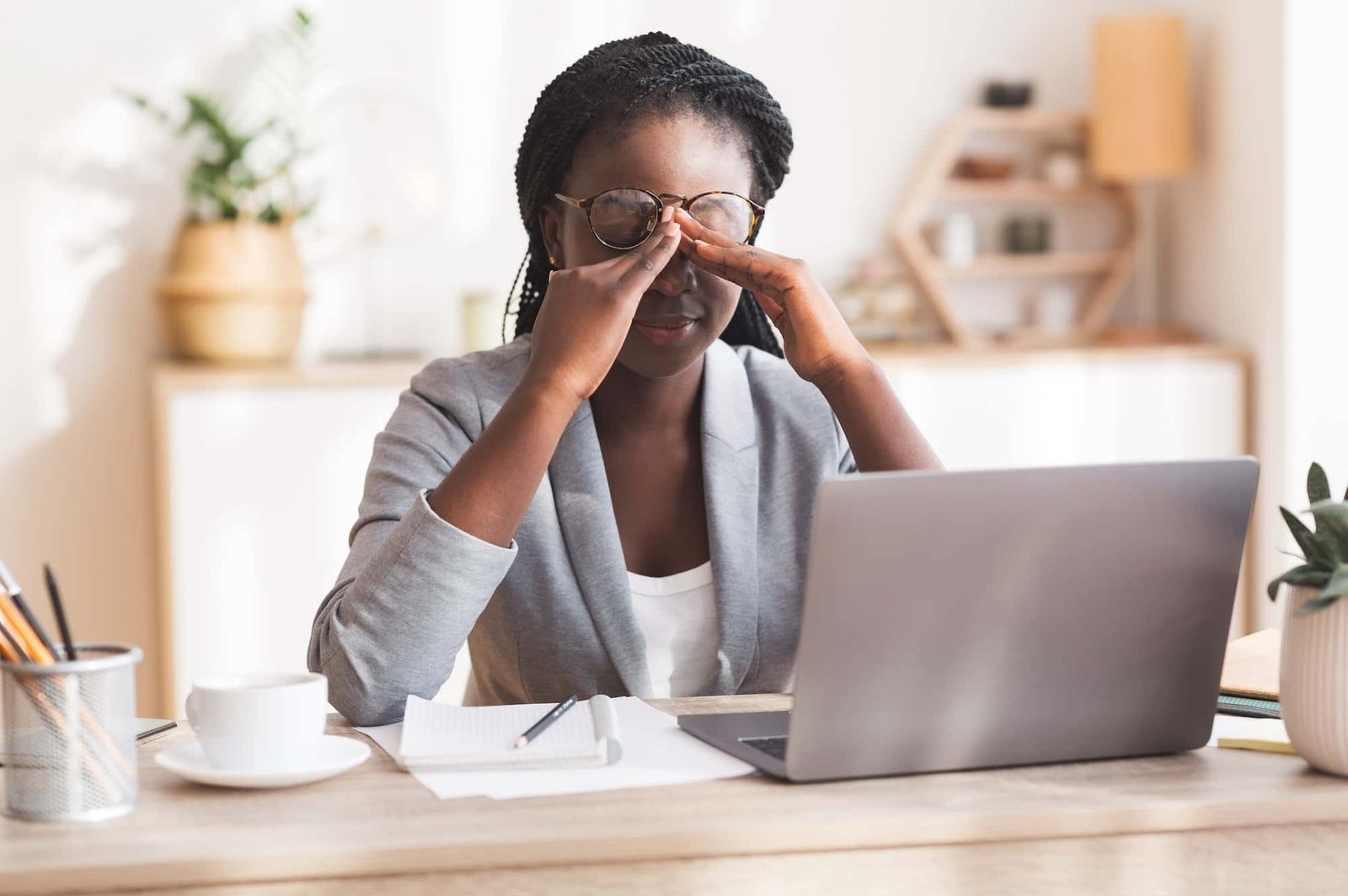 A woman sits at a desk with a laptop, rubbing her eyes as if tired or stressed from working on seo tasks. A notebook, pen, and cup are also on the desk.