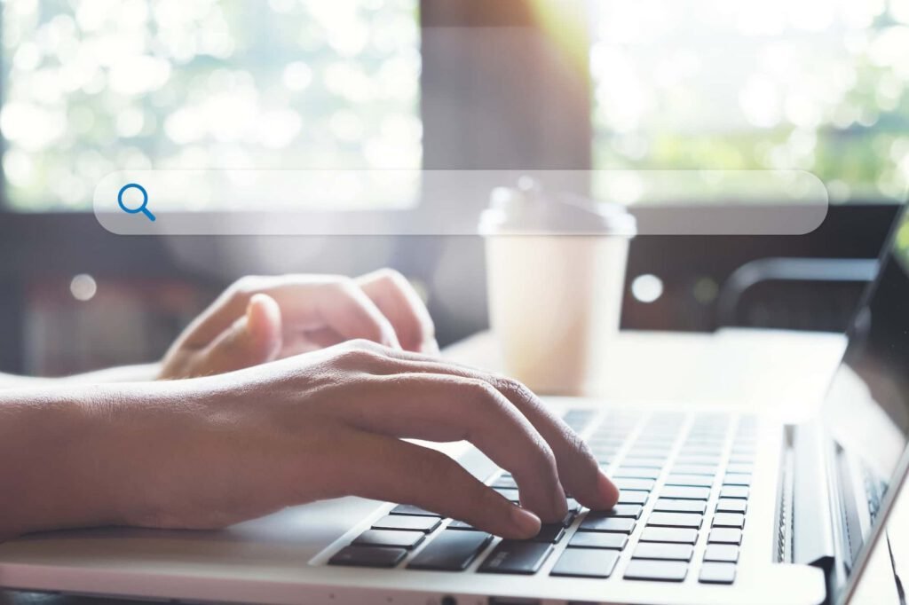 Hands typing on a laptop keyboard with a SMART search bar overlay, a paper coffee cup in the background, and sunlight streaming through a window.