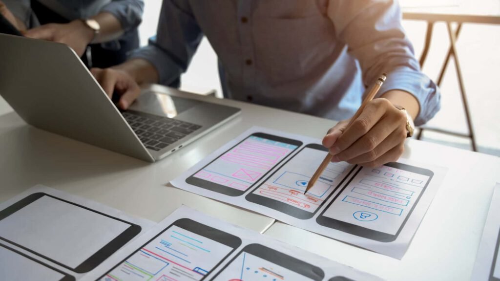 Person working on a laptop and drawing mobile app interface designs on paper prototypes at a desk, exploring website personalization features to enhance user experience.
