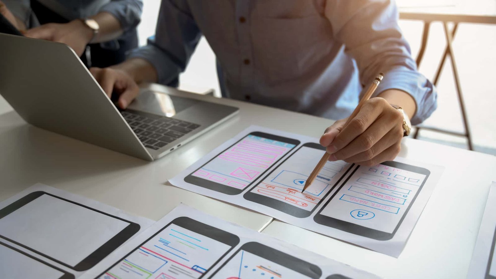 Person working on a laptop and drawing mobile app interface designs on paper prototypes at a desk, exploring website personalization features to enhance user experience.