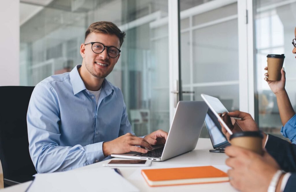 Man in glasses typing on laptop at office table with colleagues, notebooks, smartphone, and coffee cups visible; a glass-walled meeting room in the background reflects their focus on conversion-centric web design.