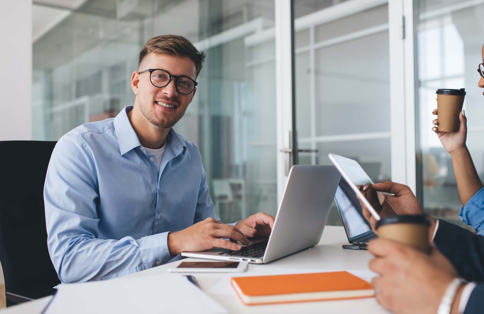 Man in glasses typing on laptop at office table with colleagues, notebooks, smartphone, and coffee cups visible; a glass-walled meeting room in the background reflects their focus on conversion-centric web design.