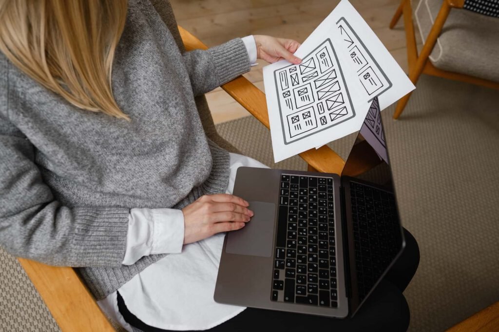 Person sitting with a laptop on their lap, holding a sheet with website wireframe sketches, suggesting work on landing page design or user interface planning.