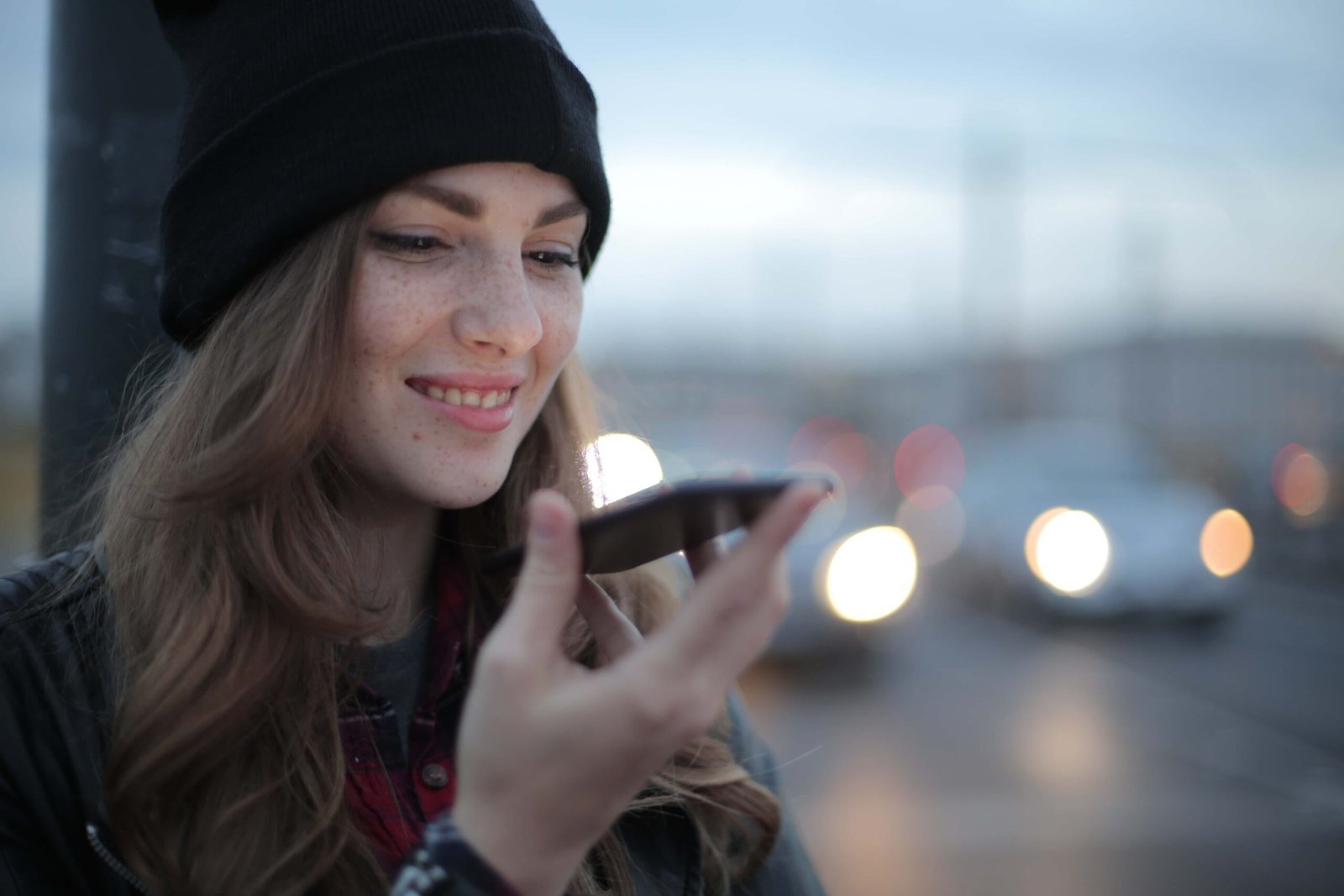 A young woman wearing a black beanie and jacket smiles while using her smartphone’s voice features outdoors in the evening.