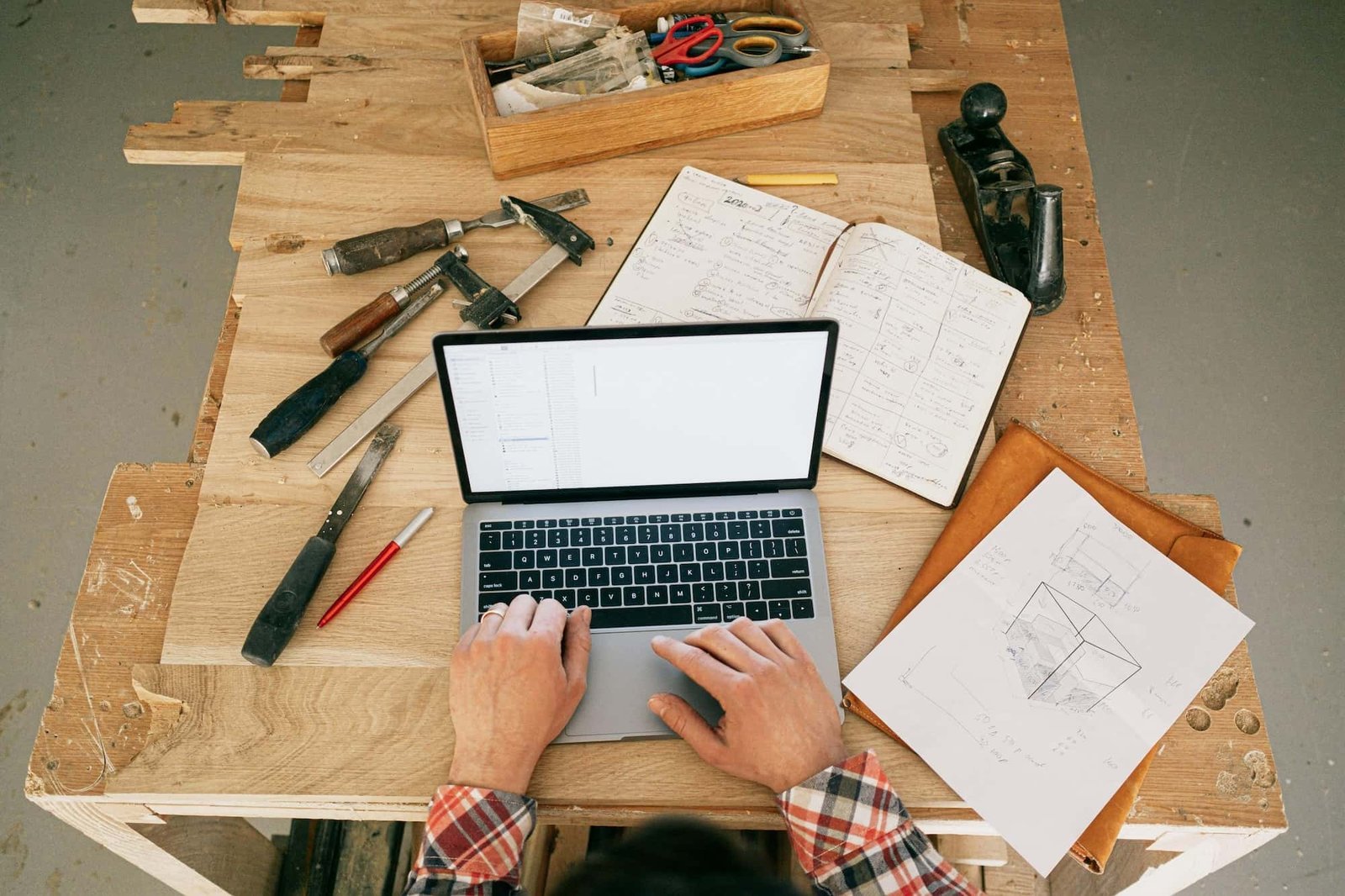Person using a laptop at a wooden workbench with construction hand tools, a notebook, a red pen, and a sheet of technical drawings.