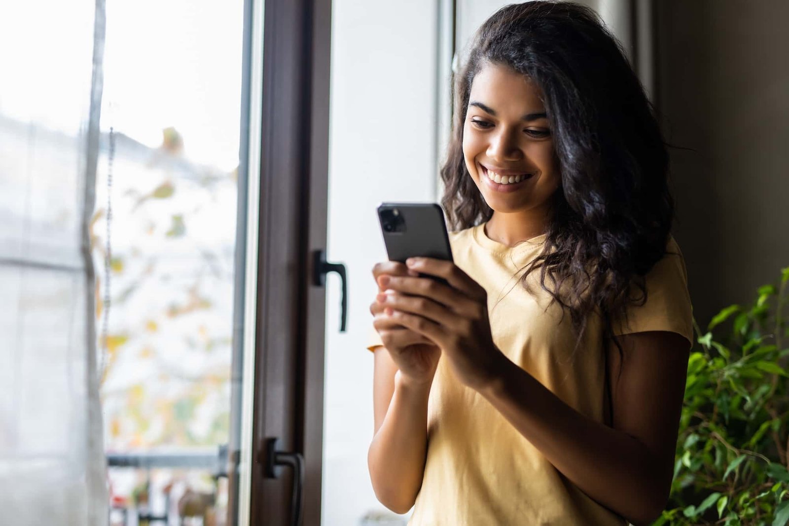 A woman stands by a window, smiling while using a smartphone. She is wearing a yellow shirt and there are green plants nearby.