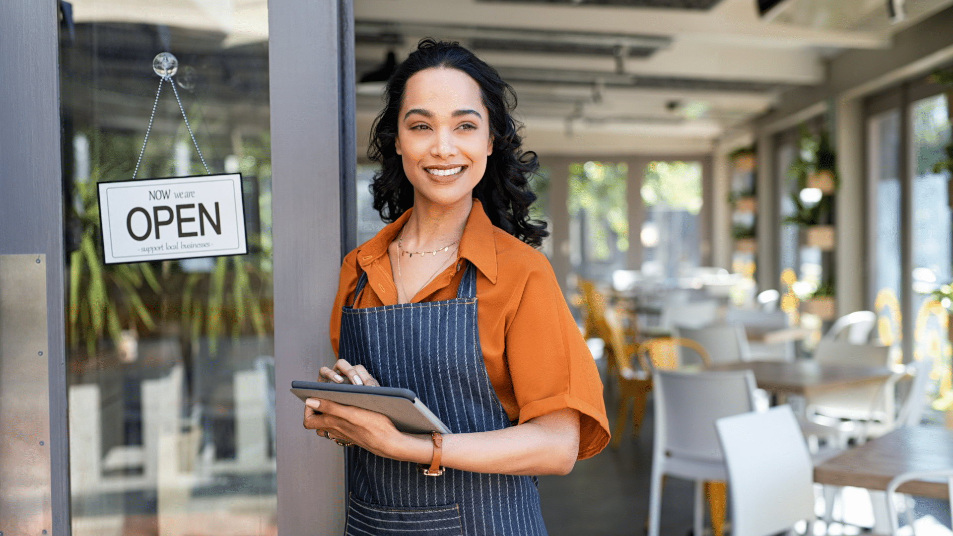 Woman wearing an apron stands at the entrance of a restaurant holding a tablet, ready to check local SEO updates. An "Open" sign hangs on the door; tables and chairs are visible inside.