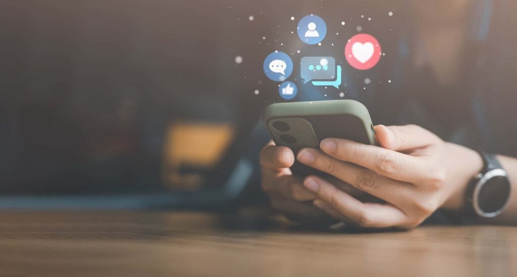 Hands holding a smartphone with floating social media icons, including like, comment, chat, and heart symbols, above the device.