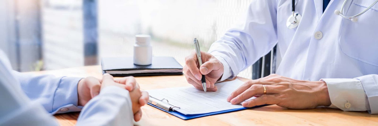 A doctor in a white coat writes content on a medical form at a desk, while a patient sits across with hands folded; a bottle of pills is on the table.