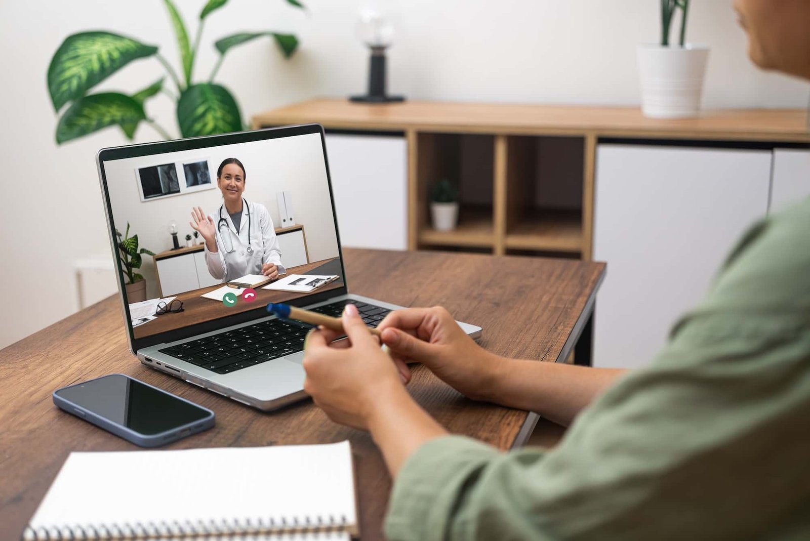 A person sits at a desk holding a pen and speaks with a doctor on a laptop during an accessible video call. A notebook and smartphone are on the table.