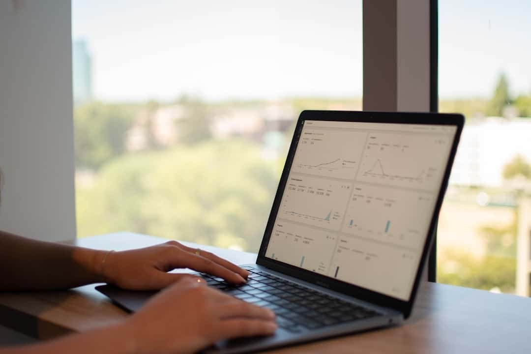 Person typing on a laptop displaying various website data charts and graphs, with a blurred outdoor view through a large window in the background.