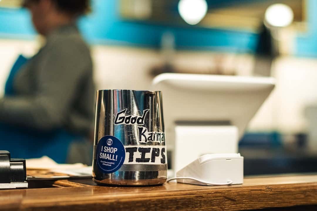 A metal tip jar labeled "Good Karma Tips" sits on a counter next to a payment terminal, subtly reminding customers that generosity—like good SEO—can boost positive vibes, with a blurred person in the background.