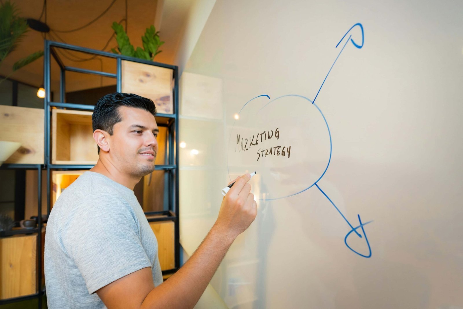 A man writes "Marketing Strategy" and diagrams on a whiteboard with blue marker, planning effective marketing approaches in a modern office setting.