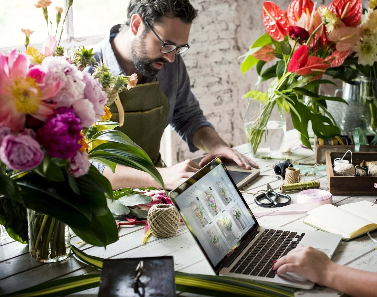 Two people work at a table covered with flowers, laptops, and floral arranging tools in a bright room, blending creative trend inspiration with web design projects.