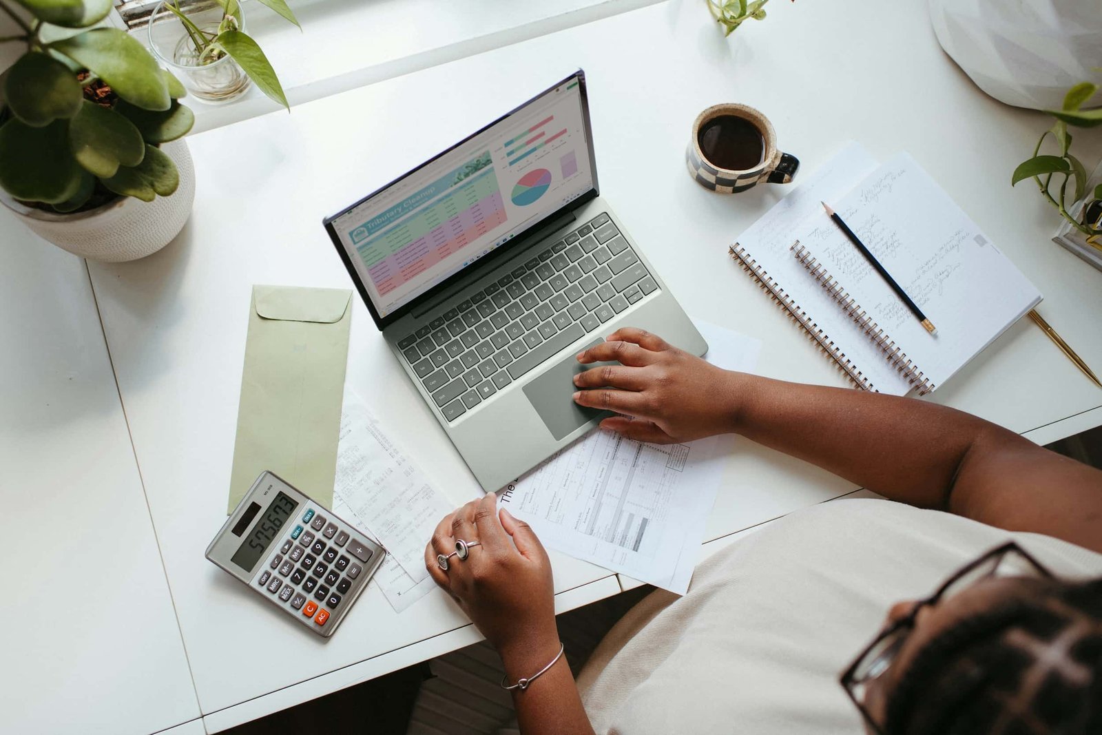 Person working at a desk with a laptop displaying SEO charts, a calculator, documents, a notepad, a pencil, an envelope, a cup of coffee, and plants nearby.