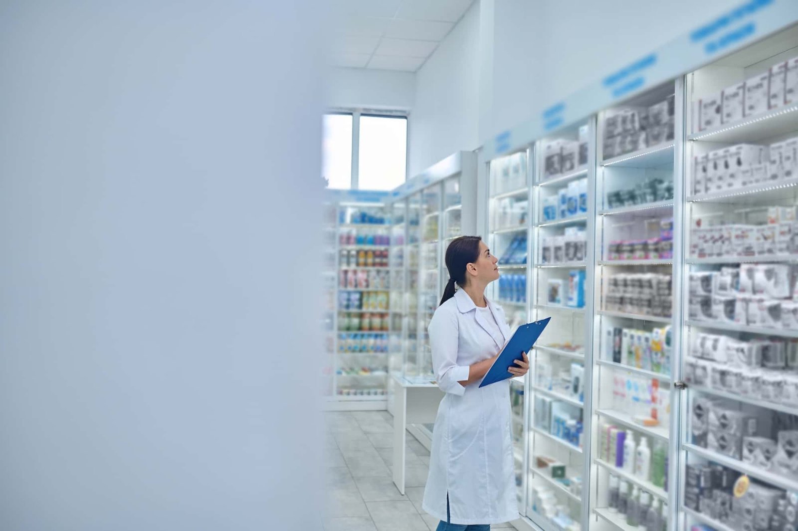 A pharmacist in a white coat holds a clipboard while inspecting shelves of medications and products in a well-lit pharmacy.
