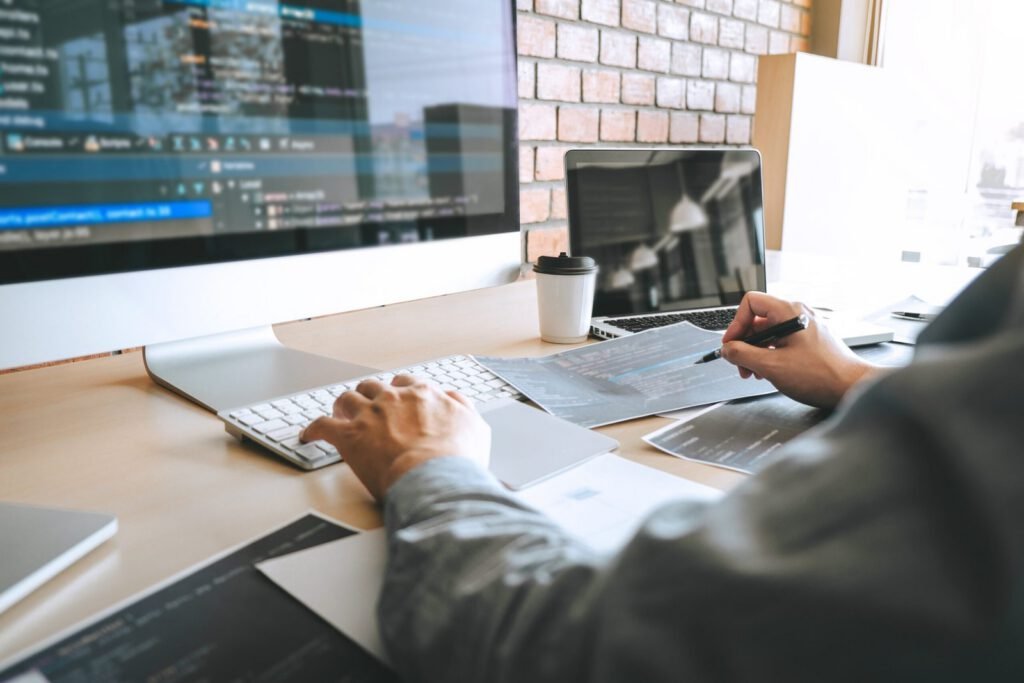 Person working at a desk with multiple monitors displaying code, using a keyboard and pen, with documents, a laptop, and a coffee cup nearby.