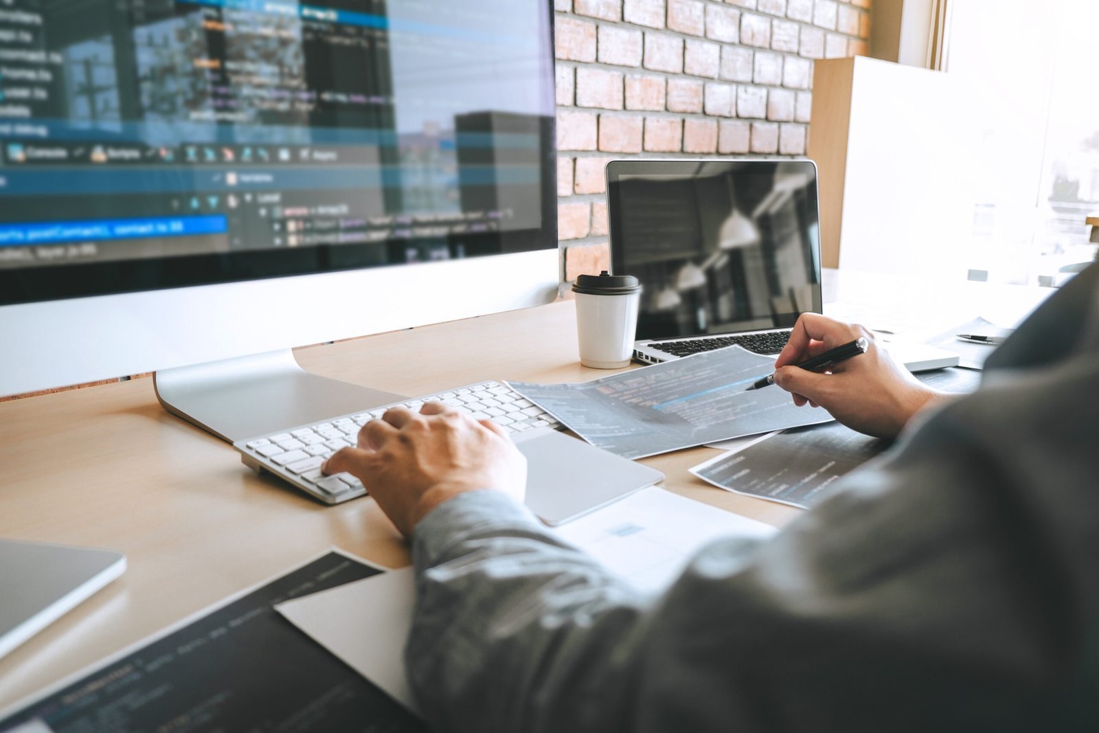 Person working at a desk with multiple monitors displaying code, using a keyboard and pen, with documents, a laptop, and a coffee cup nearby.
