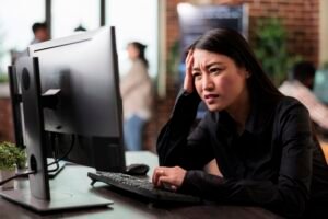 A woman sits at a desk in an office, looking frustrated while using a computer. Others are seen in the blurred background.