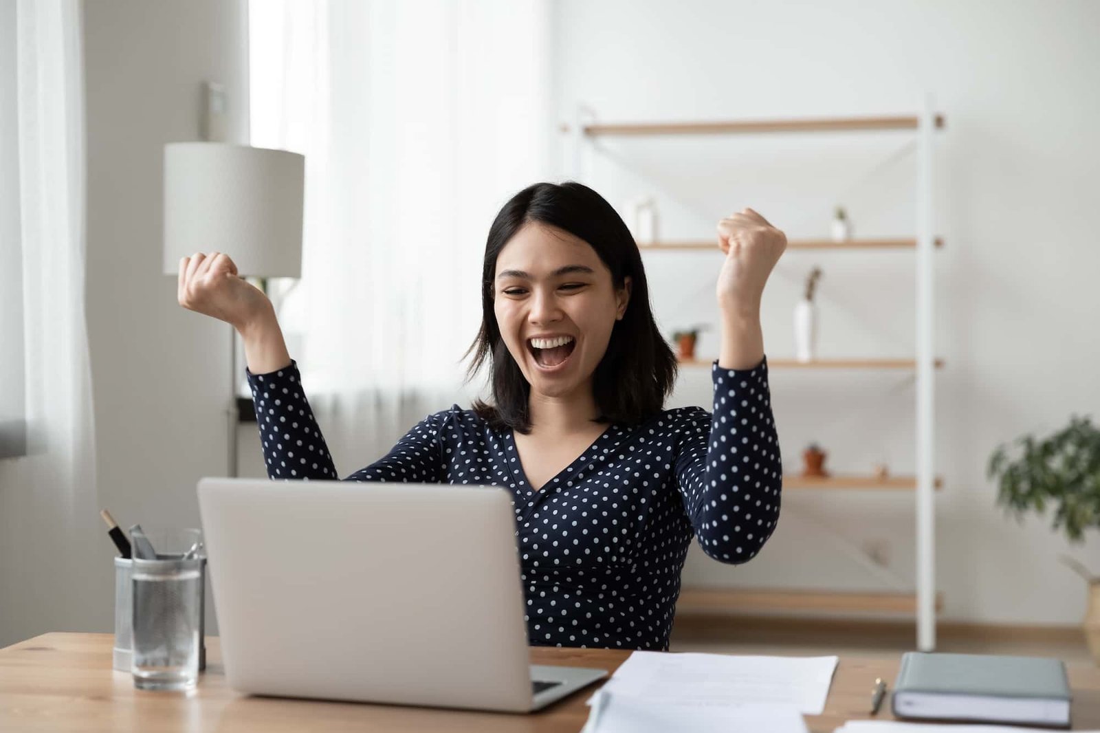 A woman sits at a desk in front of a laptop, raising her fists and smiling in excitement. Papers and a glass of water are on the desk, and shelves are in the background.