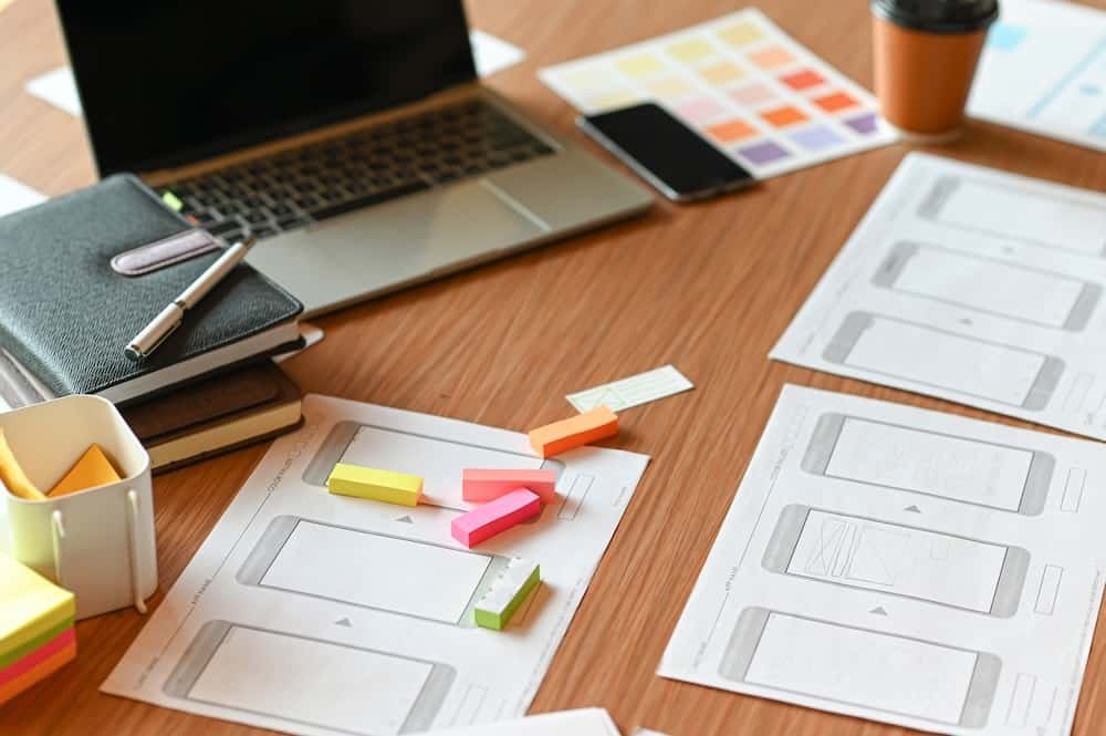 A desk with wireframe sketches, colorful sticky notes, a laptop showcasing web design ideas, a phone, a notebook with a pen for brainstorming SEO strategies, and a coffee cup.